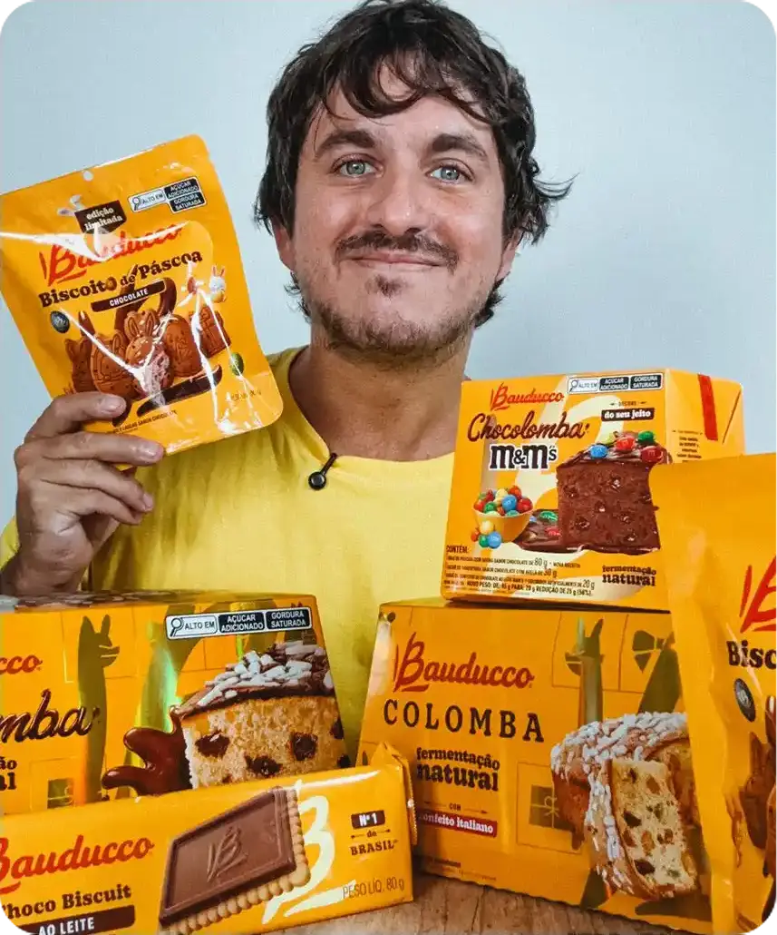Homem sorrindo e segurando produtos Bauducco, incluindo Biscoito de Páscoa, Choco Biscuit e Colomba. Tom alegre, otimista e promocional.