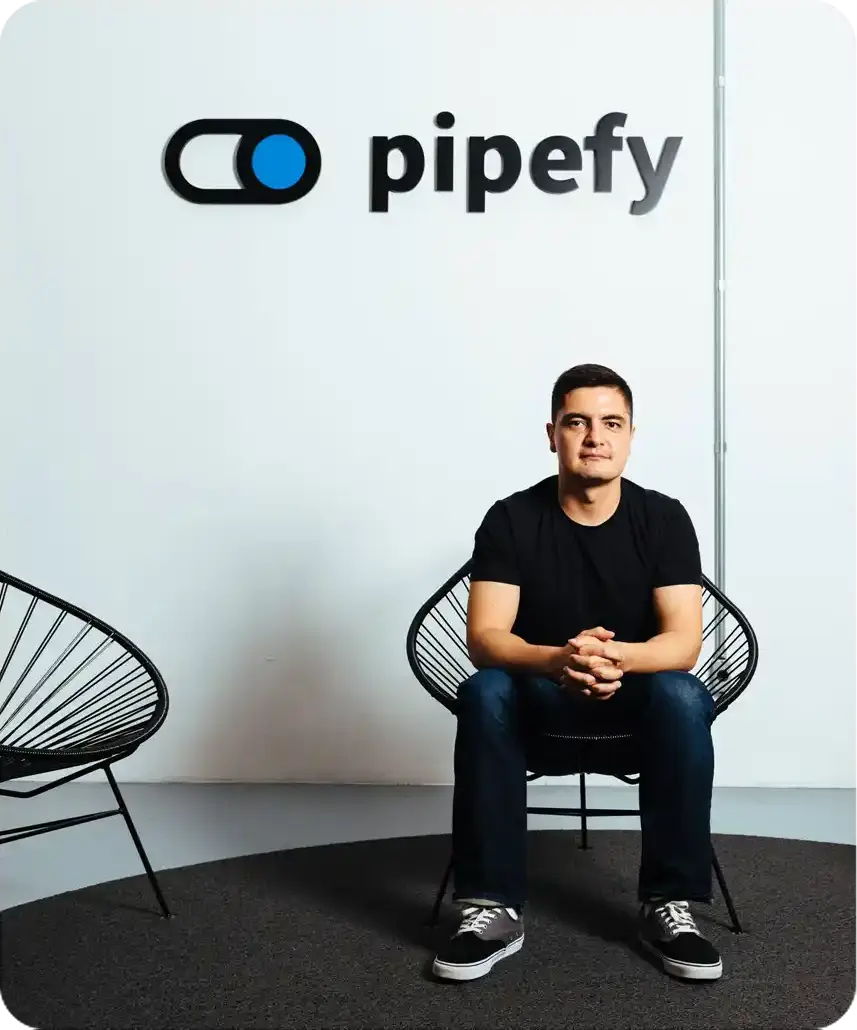 Um homem de camiseta preta senta-se com confiança em uma cadeira em um escritório moderno com o logotipo 'pipefy' na parede; ambiente minimalista e profissional.