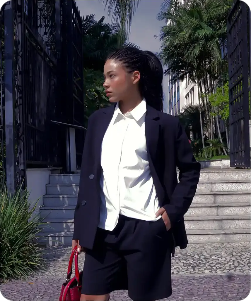 Uma mulher com um elegante blazer preto e shorts, segurando uma bolsa vermelha, está de pé com confiança perto de uma entrada com portão, cercada por vegetação exuberante e degraus.
